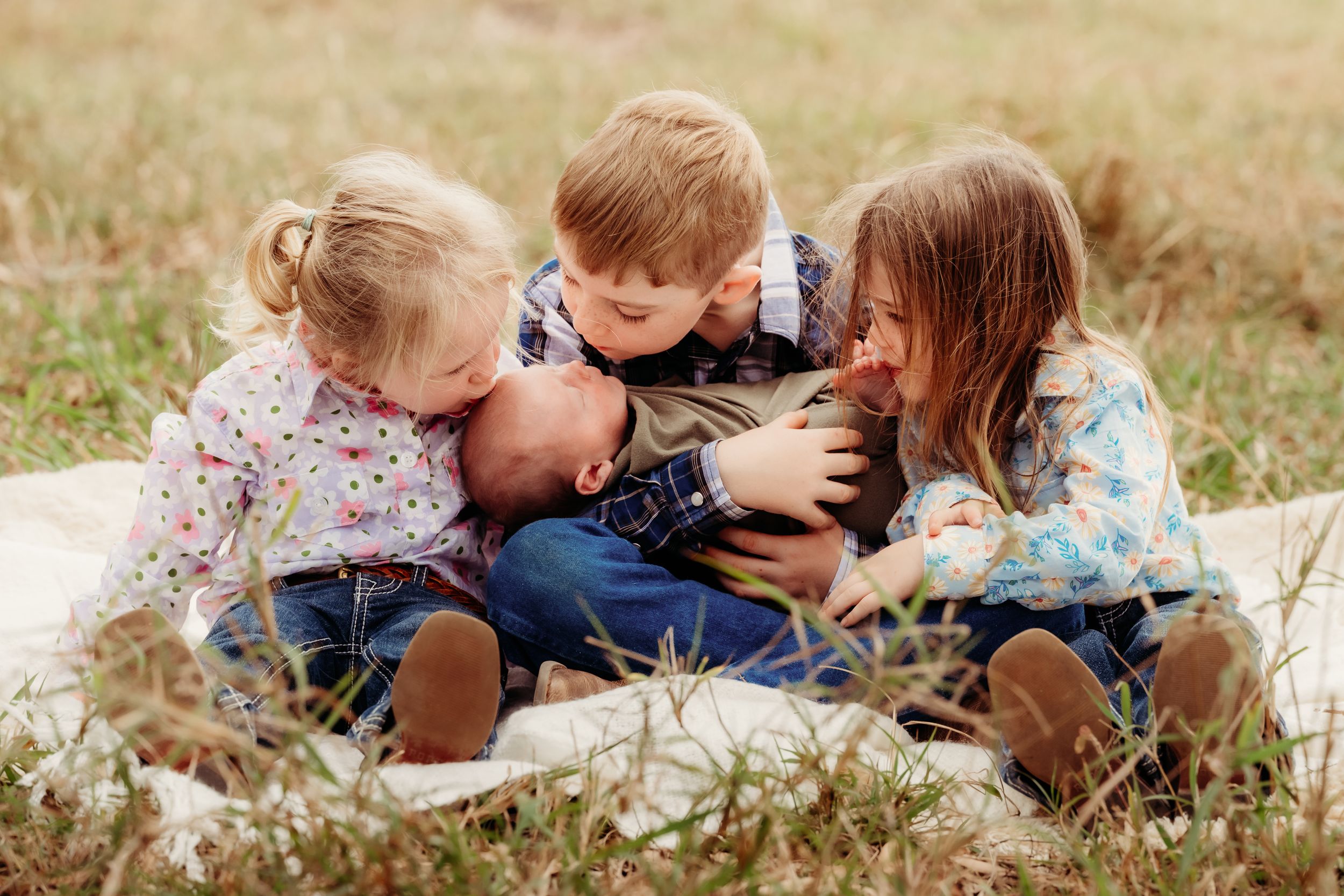 Four young children in jeans and plaid shirts huddle together on a white blanket in a grassy field, cuddling a baby.