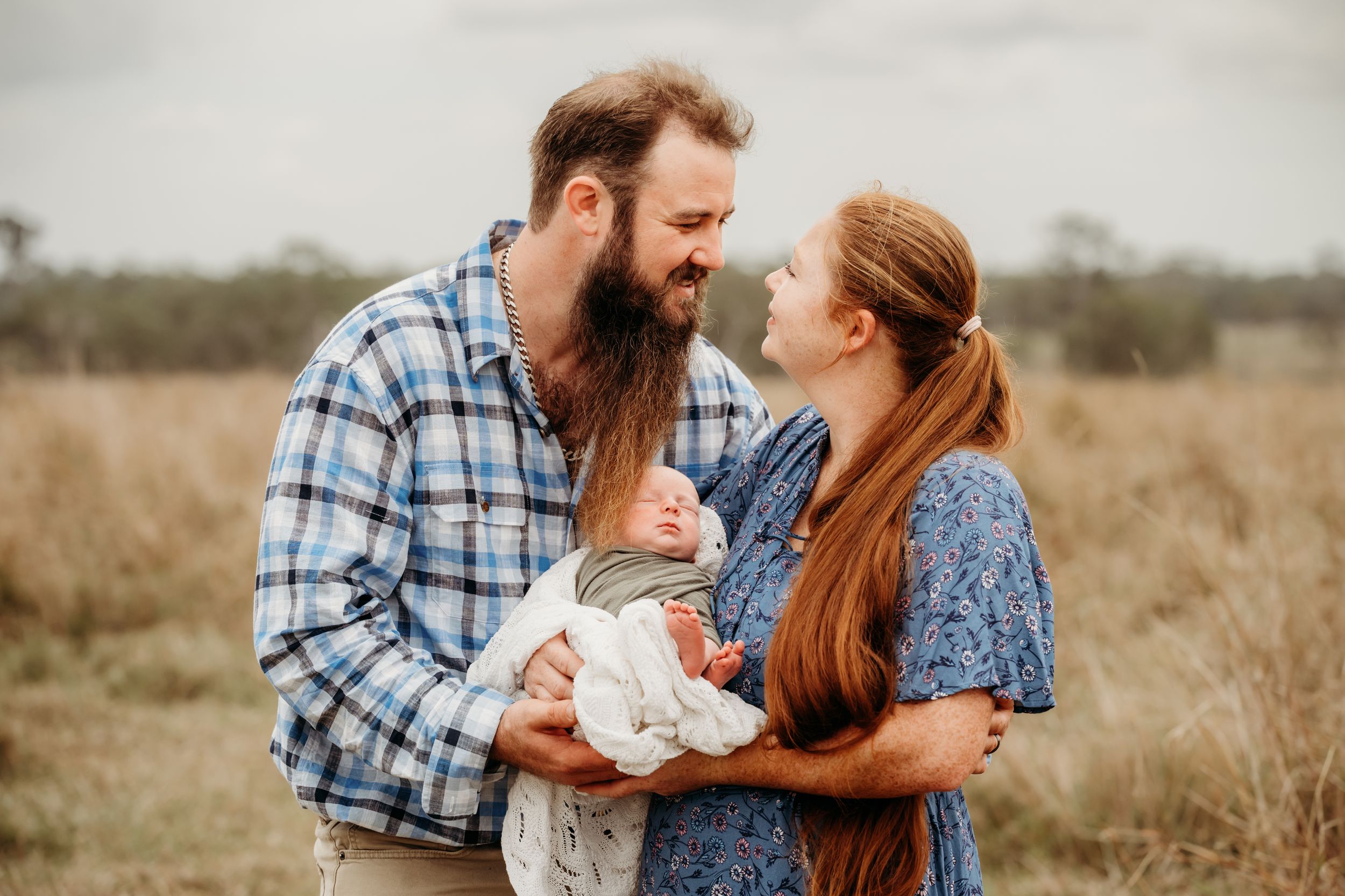 Bearded man in a blue plaid shirt and woman with long auburn hair smile at their newborn baby in a golden grassy field.