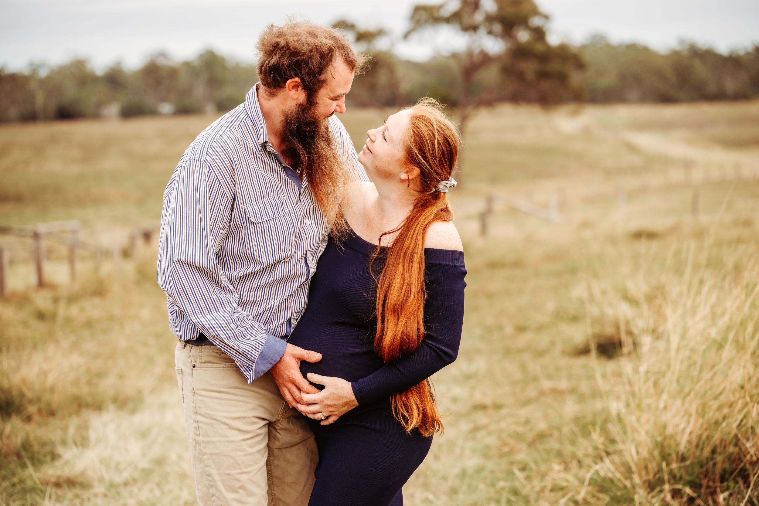 A couple shares a tender moment in a field, the woman visibly pregnant in a navy dress, her red hair flowing.