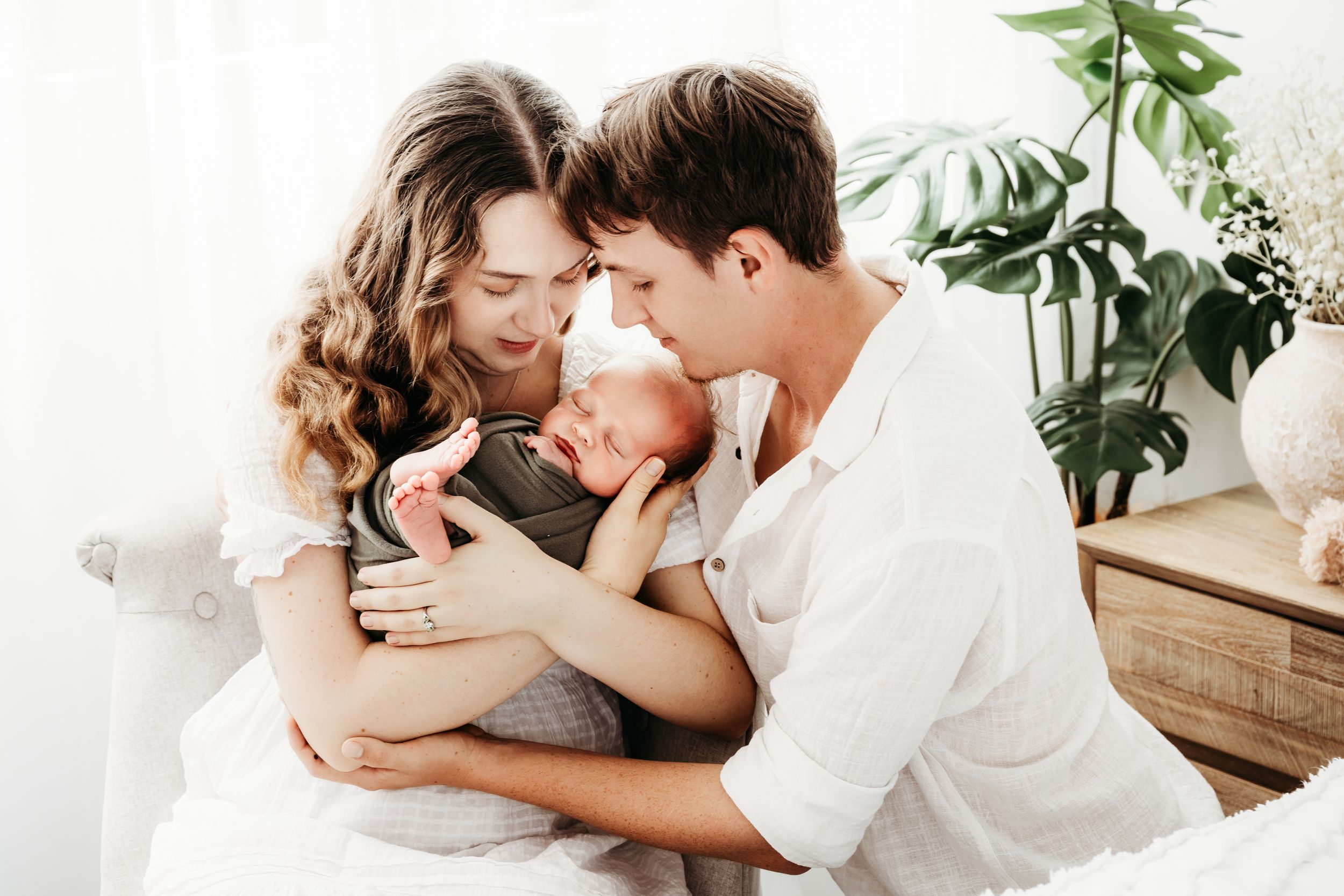 Intimate family moment with new parents cradling their newborn baby near a houseplant.