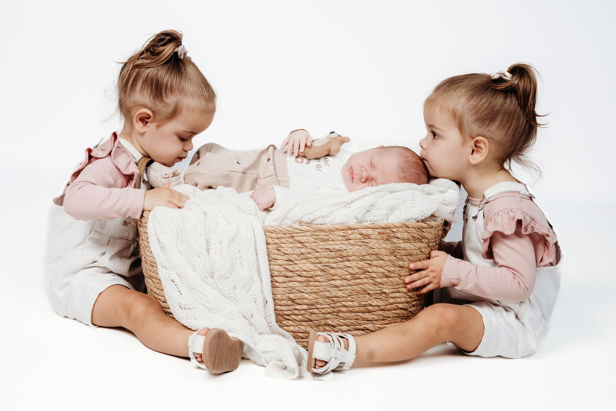Two toddler girls sitting beside a wicker basket holding a sleeping newborn baby, on a white background.