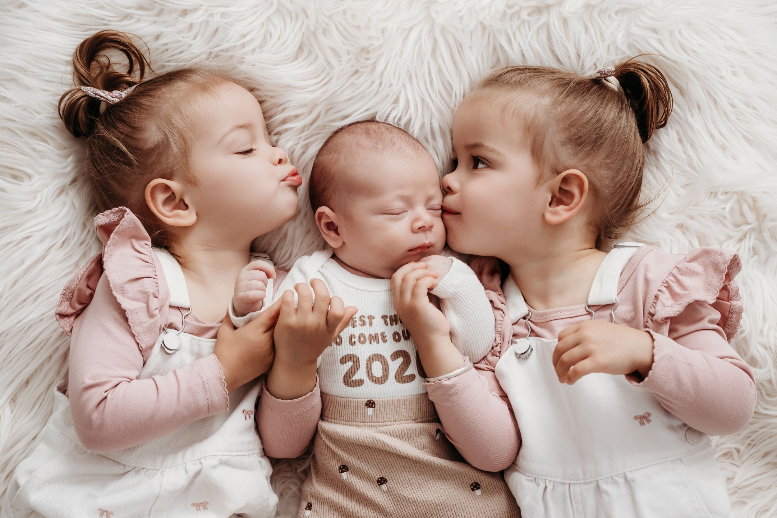Two toddler girls in pink outfits kiss a sleeping newborn baby wearing a 2020 onesie on a white fluffy blanket.