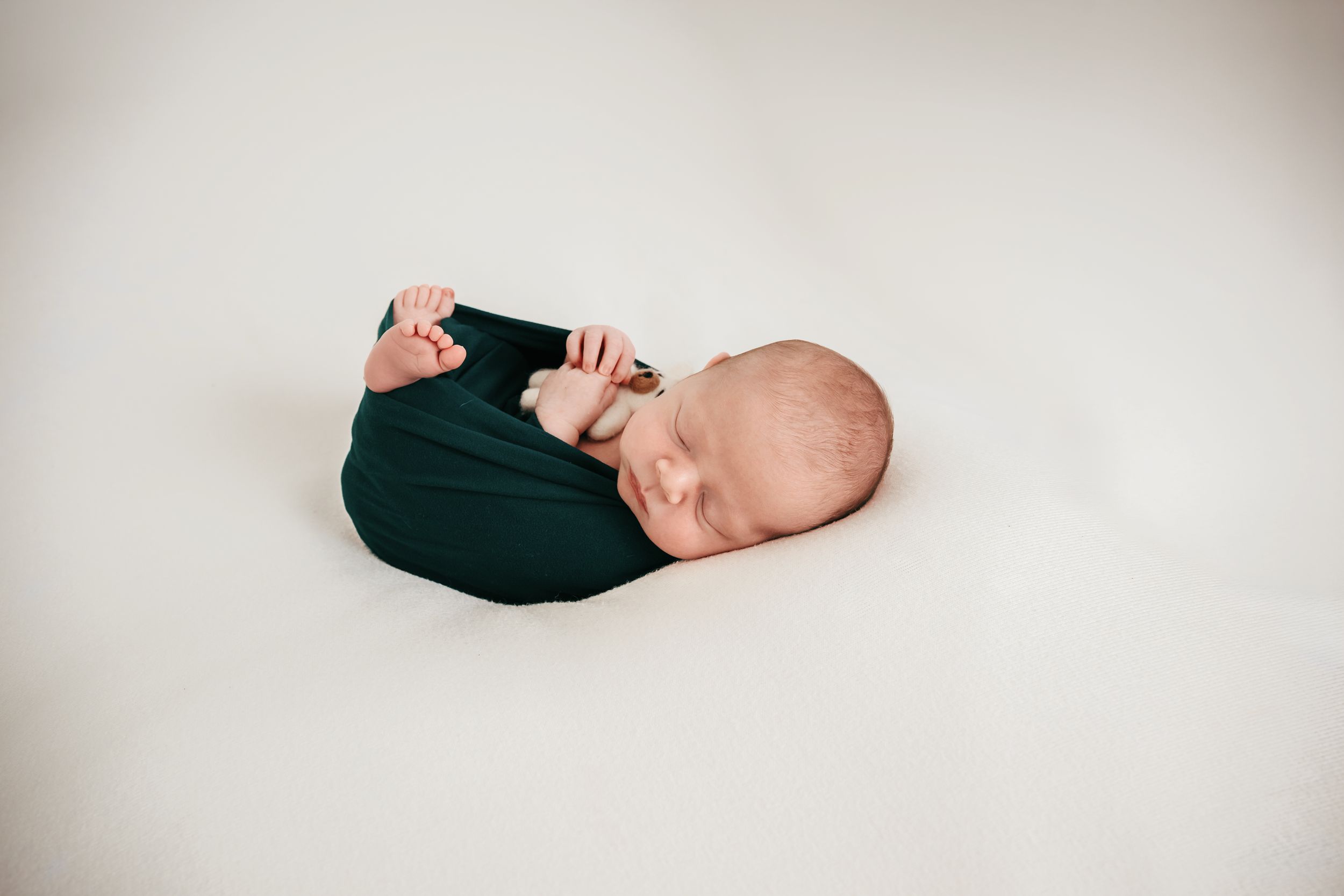 Newborn baby swaddled in a dark green wrap, sleeping peacefully on a white background in a professional photo.