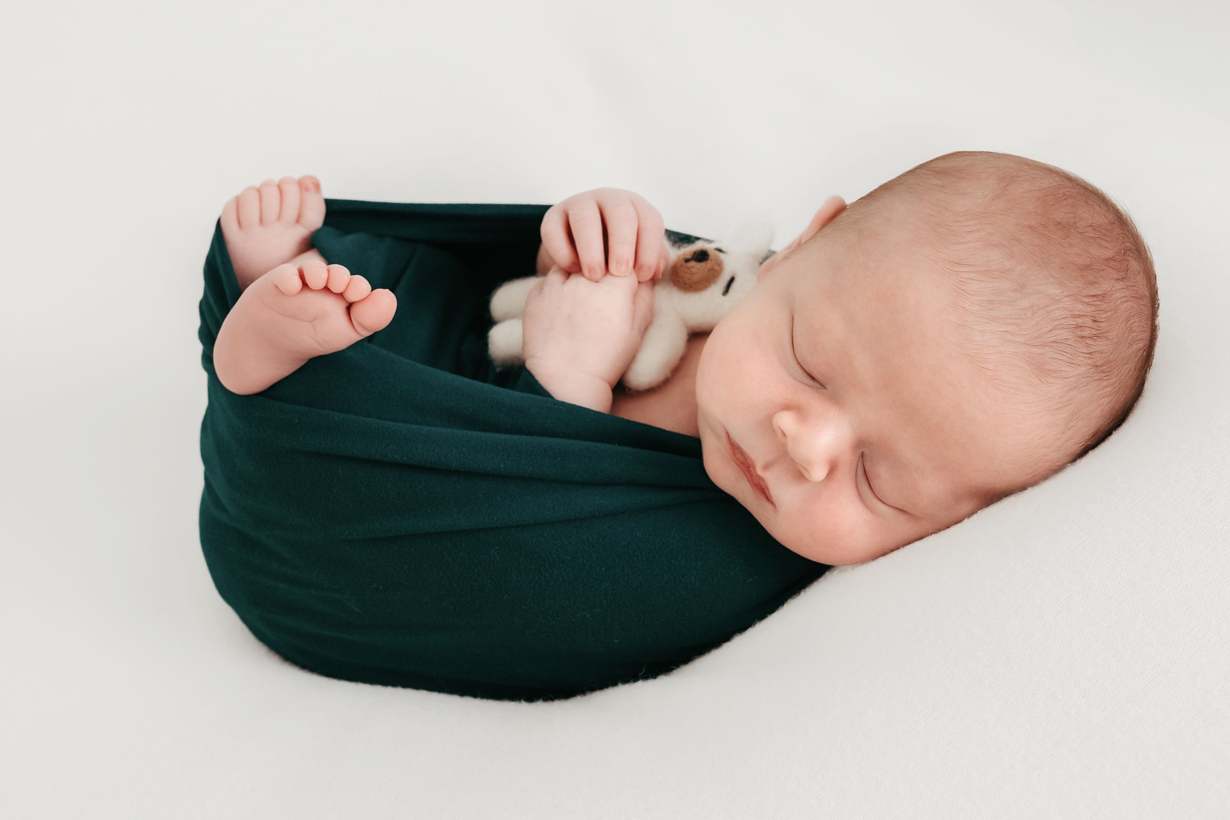 Sleeping newborn baby wrapped in a dark green swaddle blanket on a white background.