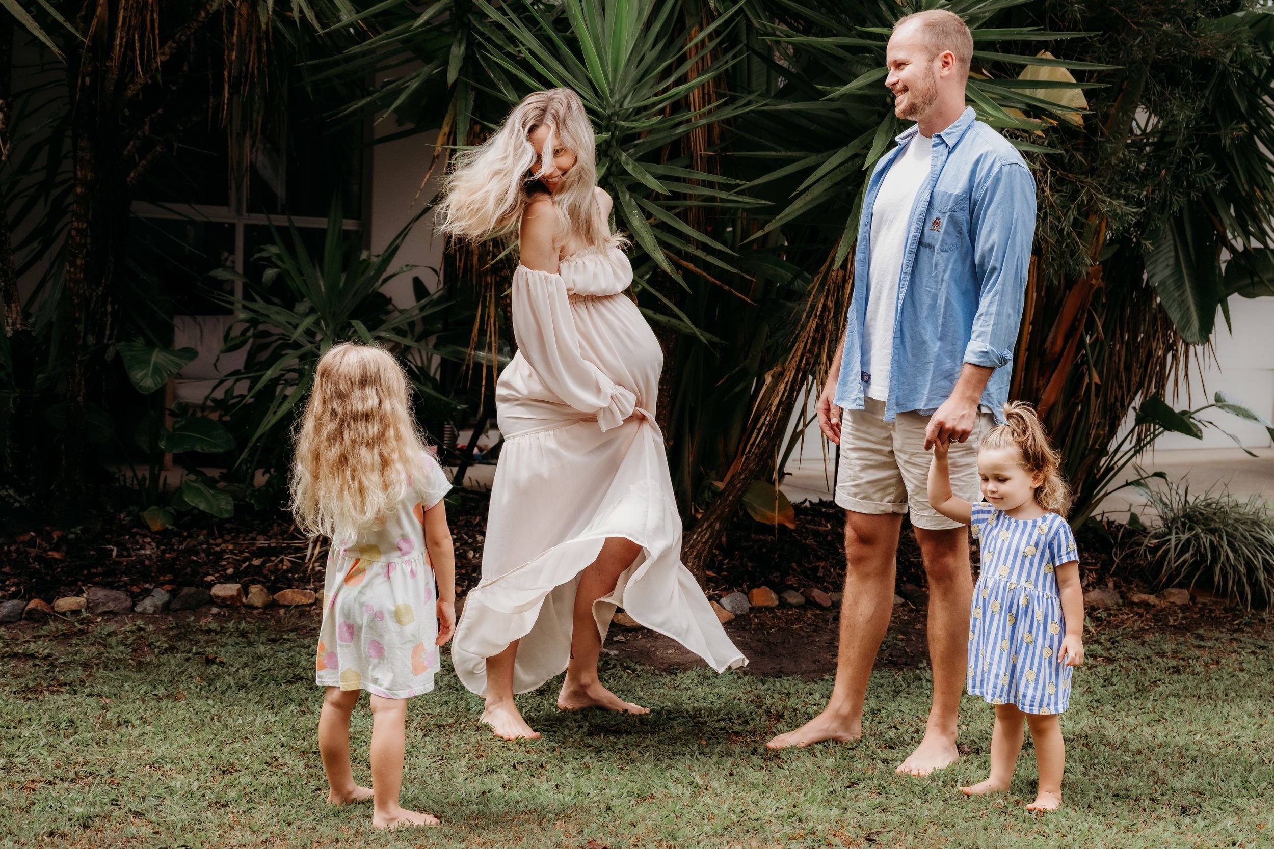 Happy barefoot family with two young girls dancing outdoors on green grass with tropical trees in background.