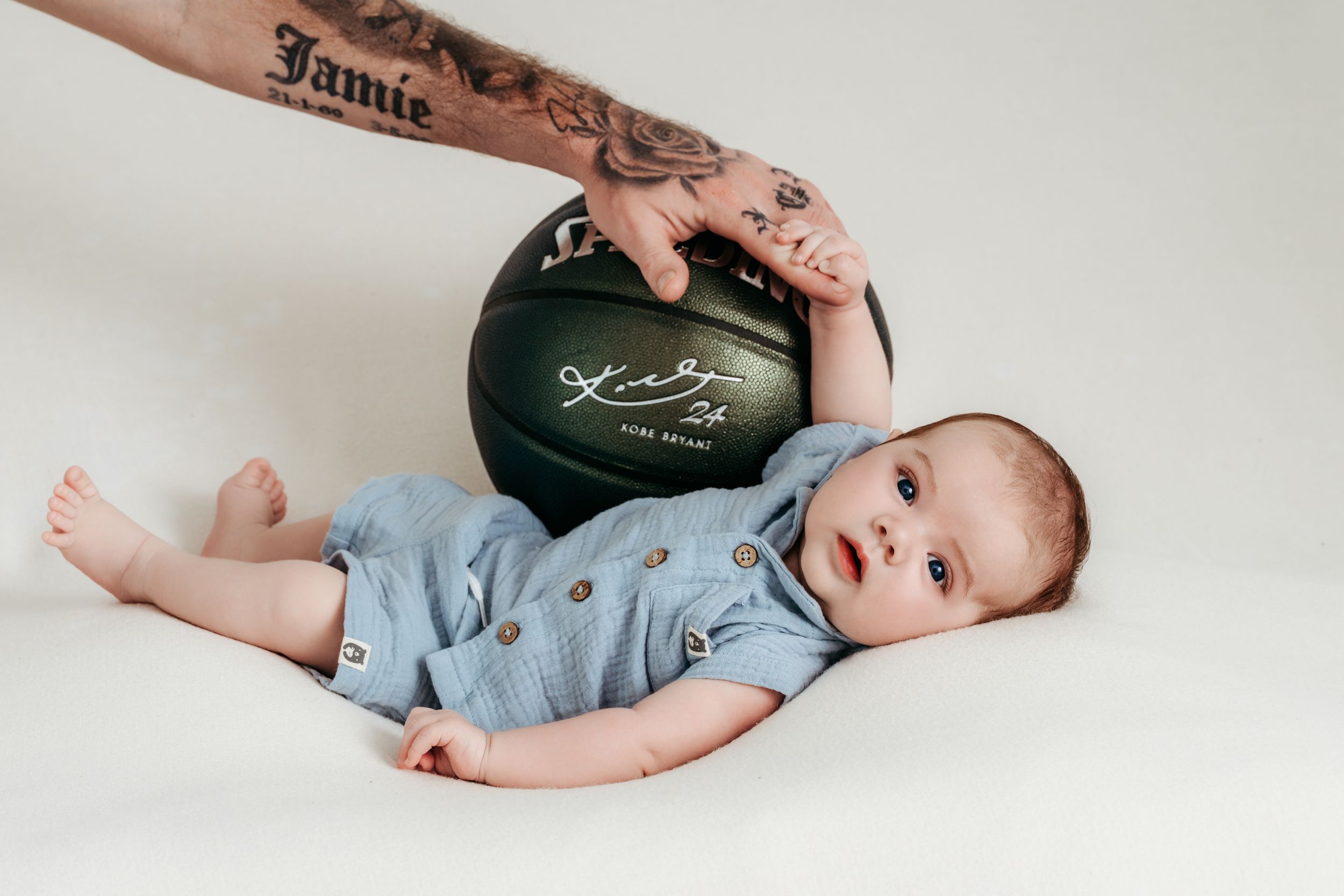 Baby in blue romper lying next to a dark green basketball held by a tattooed hand.