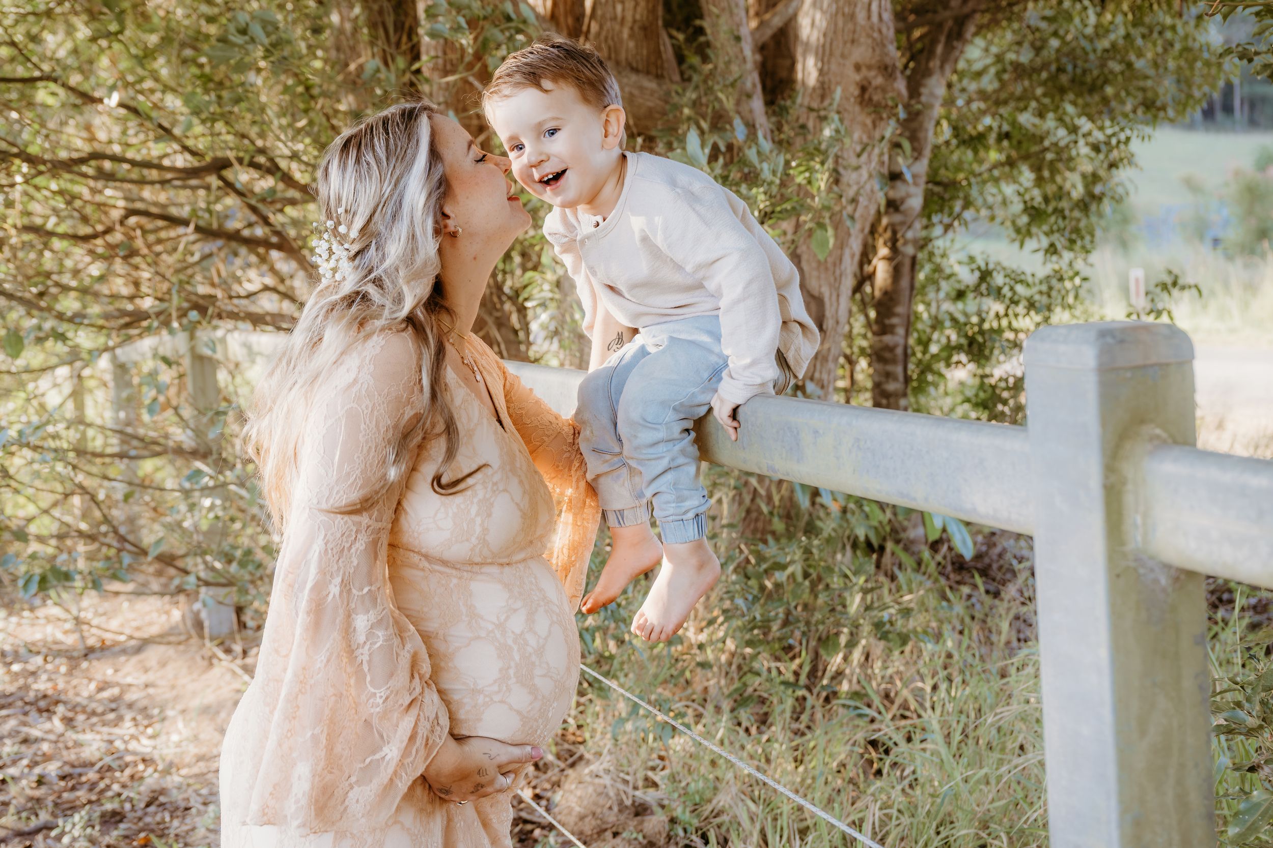 Pregnant woman in beige dress laughing with toddler boy sitting on white fence outdoors.