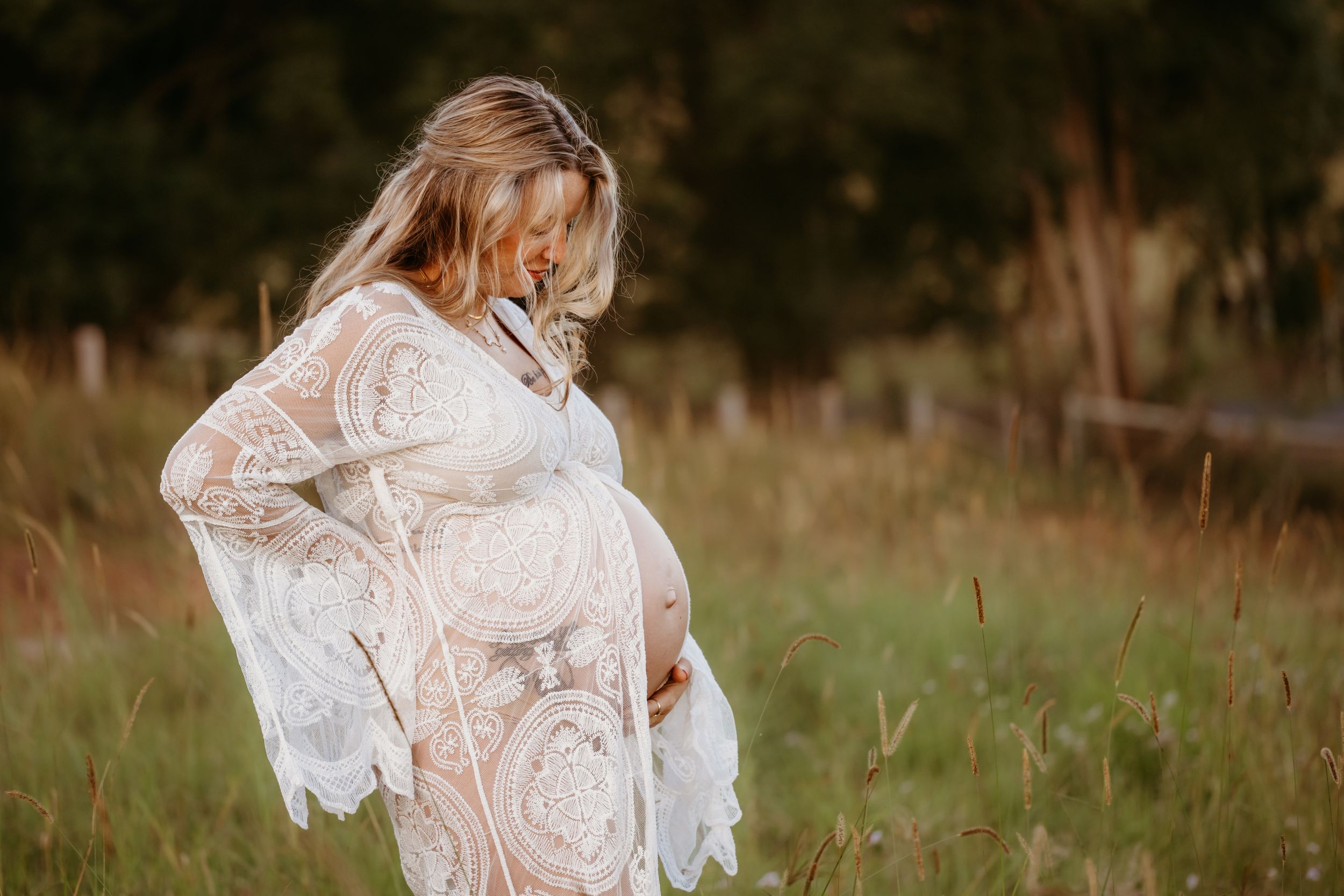 Pregnant woman in white lace boho dress cradling baby bump in sunlit meadow during golden hour maternity photoshoot.
