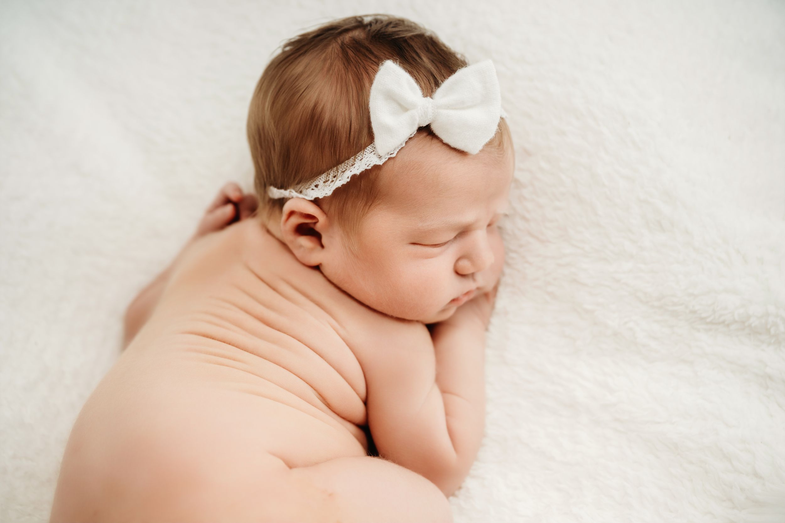Peaceful newborn baby girl sleeping on white fluffy blanket wearing a delicate white bow headband.