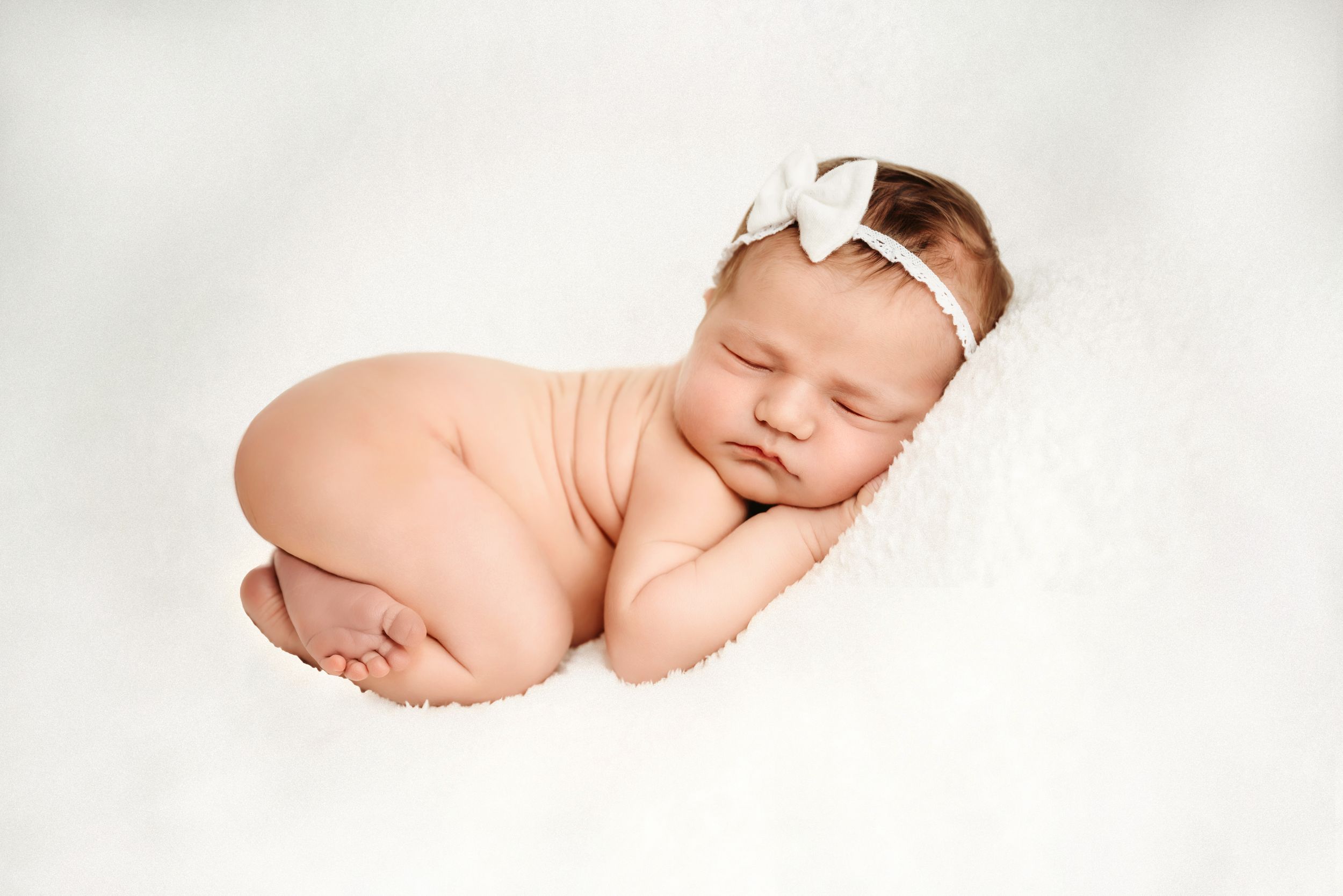 Peaceful newborn baby girl sleeping curled up on white fluffy blanket wearing a delicate white bow headband.