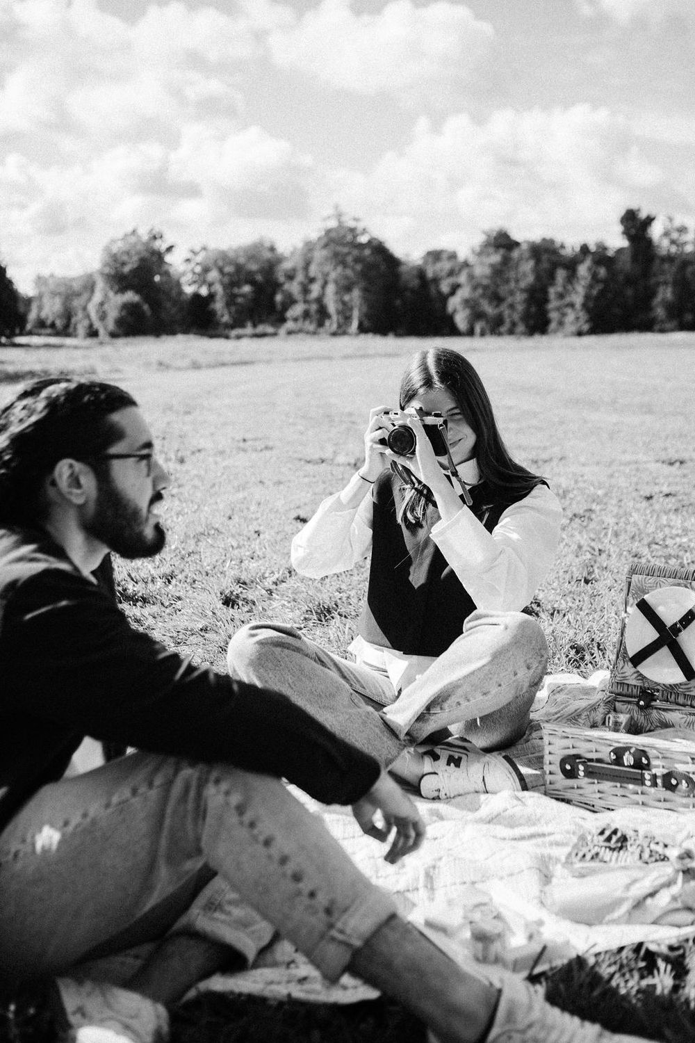 Black and white photo of a couple enjoying a picnic in a field, woman photographing man with a vintage film camera.
