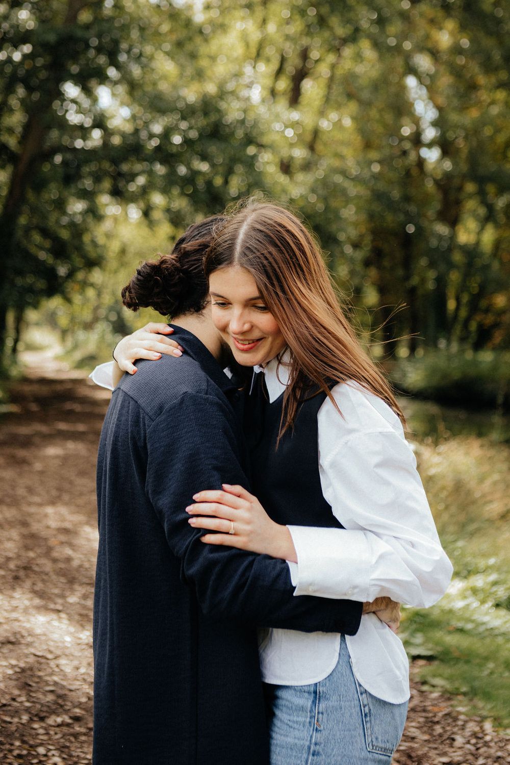 Young couple sharing a tender hug on a tree-lined forest path, woman smiling in white shirt and jeans.