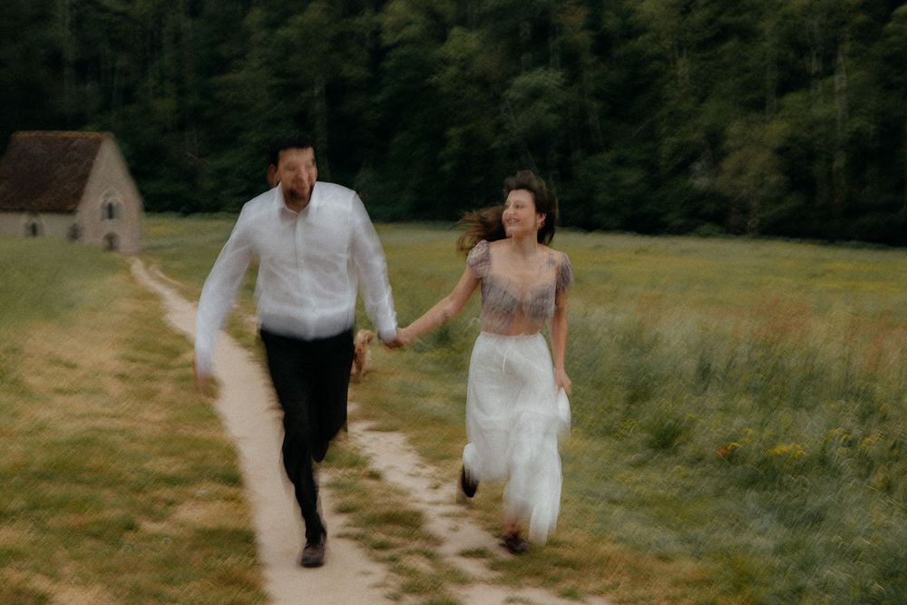 Couple in wedding attire running hand-in-hand along a dirt path through a lush green meadow.