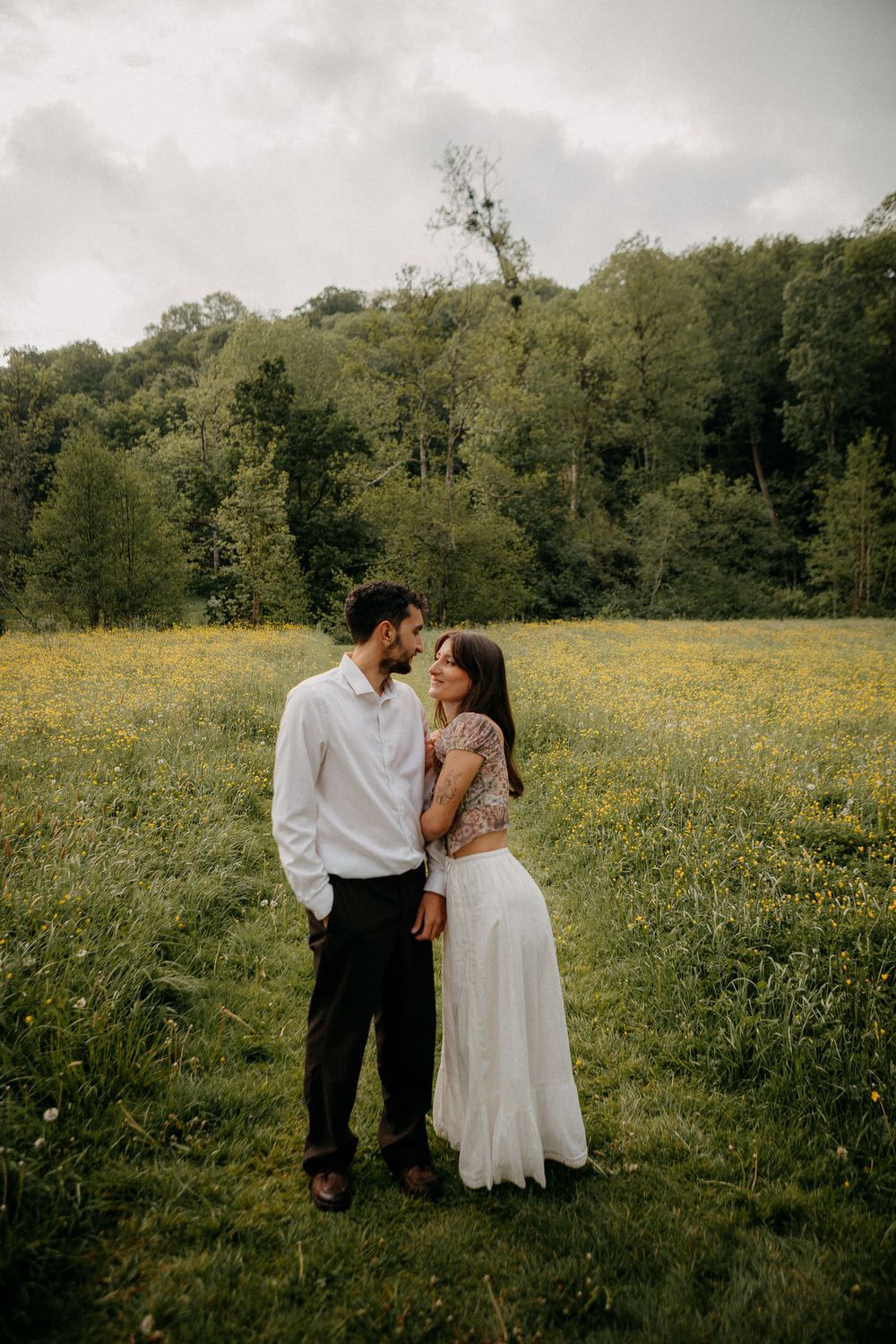 Couple sharing a romantic moment in a wildflower meadow, surrounded by lush green trees under a cloudy sky.