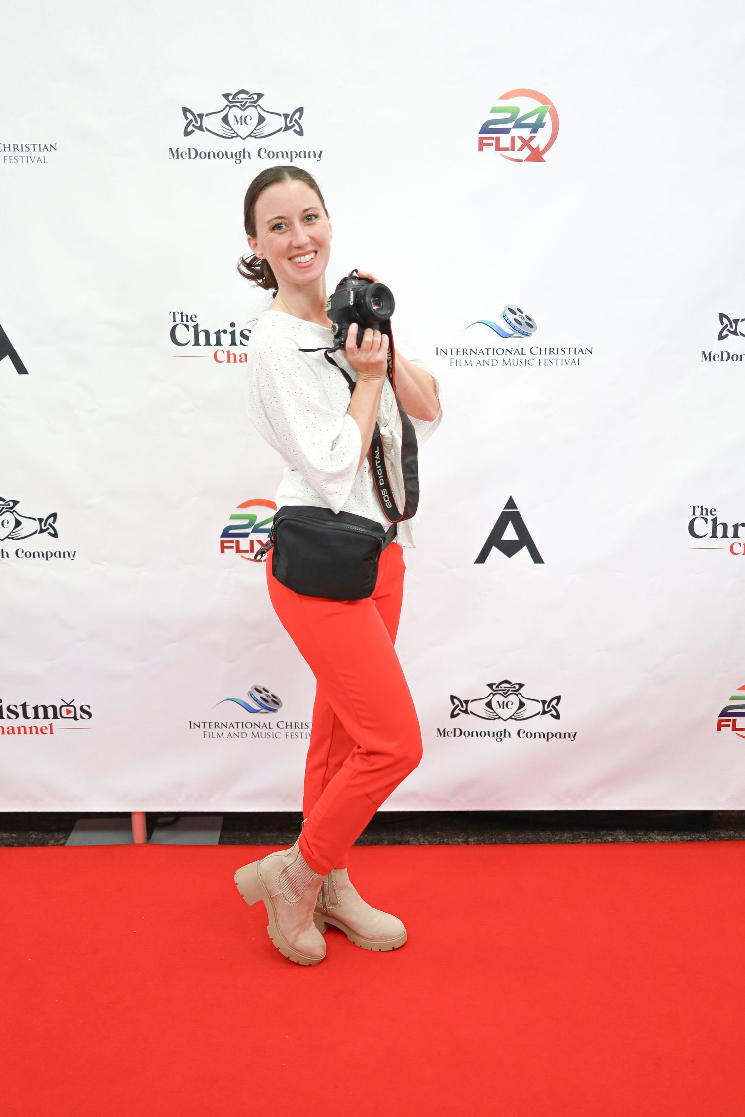 A woman in a white top and red pants poses playfully on a red carpet at a branded event backdrop.