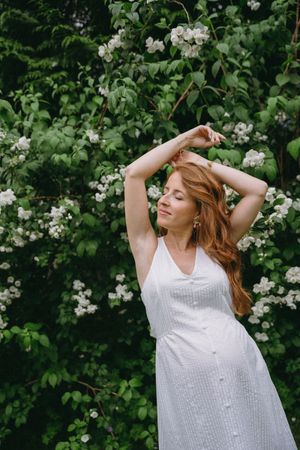 Woman in white dress poses elegantly against flowering bushes in a garden setting.