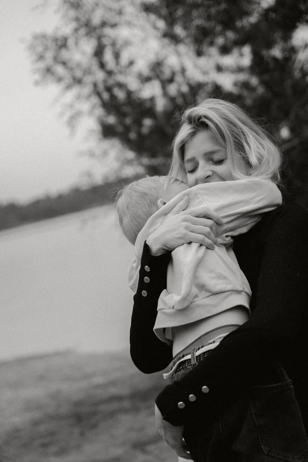 Parent embraces young child in tender black and white portrait by waterfront.