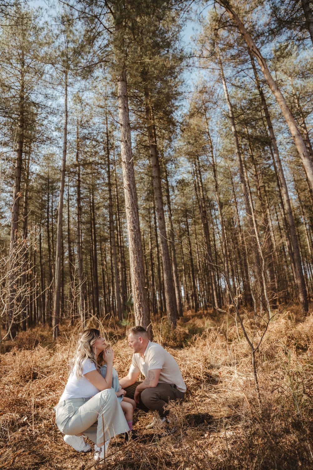 Couple sits together in sunlit pine forest with fallen needles.