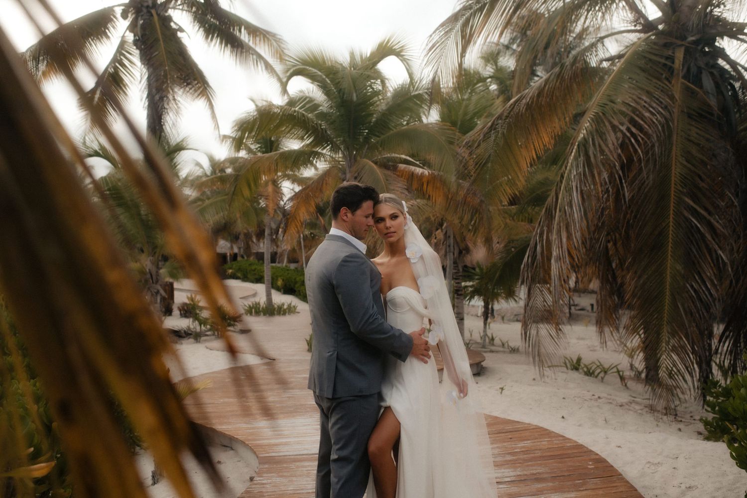 A couple shares a romantic moment on a beachside walkway surrounded by tropical palm trees at sunset.