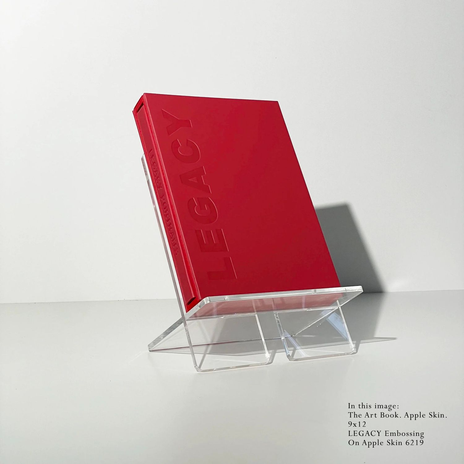 Red hardcover Legacy art book displayed on a clear acrylic stand against white background.
