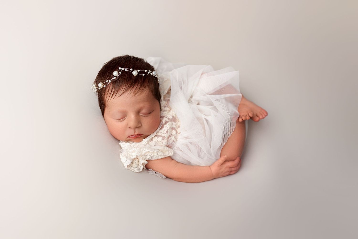 Sleeping newborn baby in grey outfit and white bonnet on light backdrop.