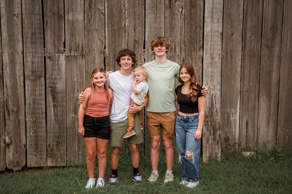 Five siblings including a toddler posing together in front of a weathered wooden fence outdoors.
