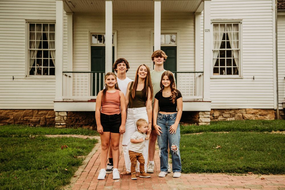 Six siblings with a toddler posing in front of a charming white historic house on a brick path.