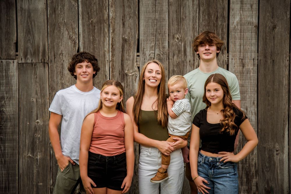 Six siblings posing together in front of a rustic wooden barn wall for a family portrait.
