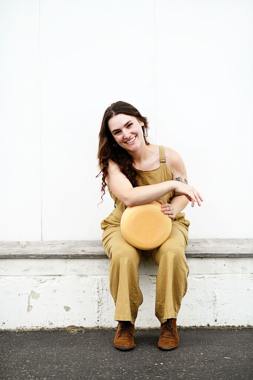 Woman in mustard overalls sits casually with round cheese wheel against white wall.