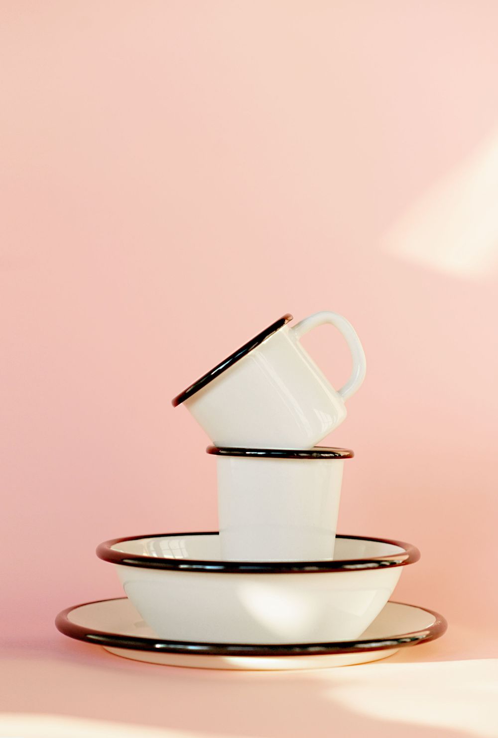 Minimalist stack of white enamel cups and plates with black trim against pink background.