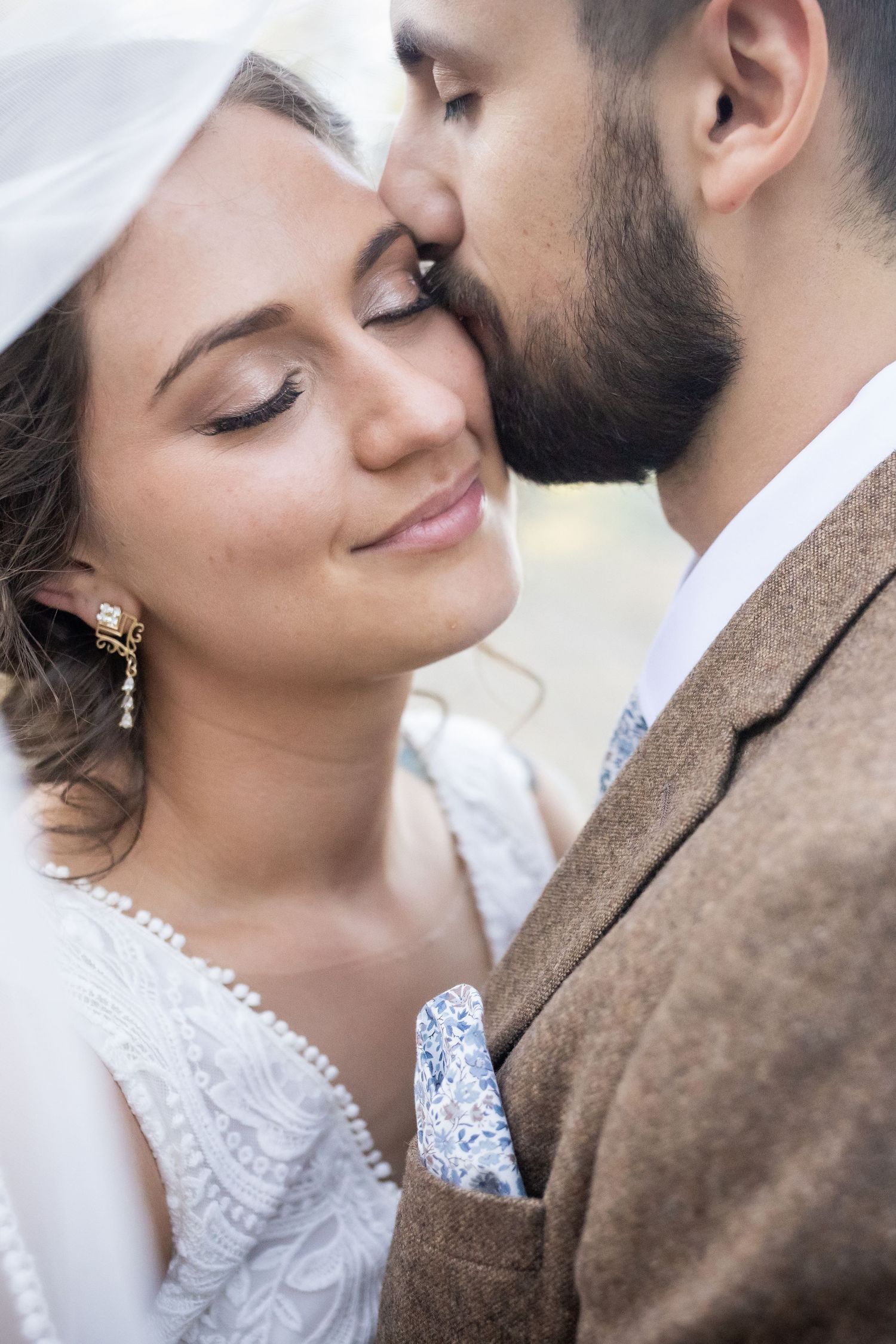 Groom gently kissing bride on cheek as she smiles softly in romantic wedding portrait.