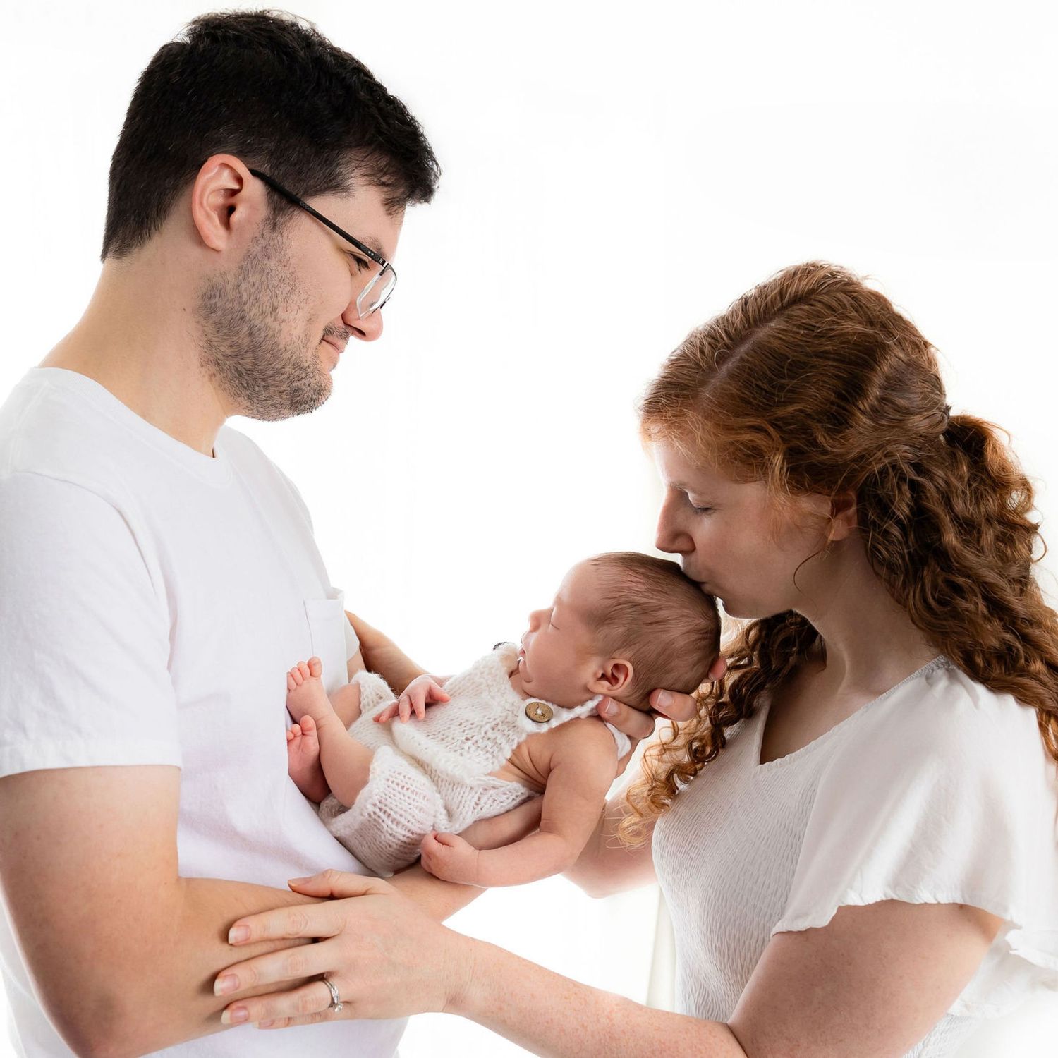 Parents in white attire share a tender moment with their newborn baby against a light background.
