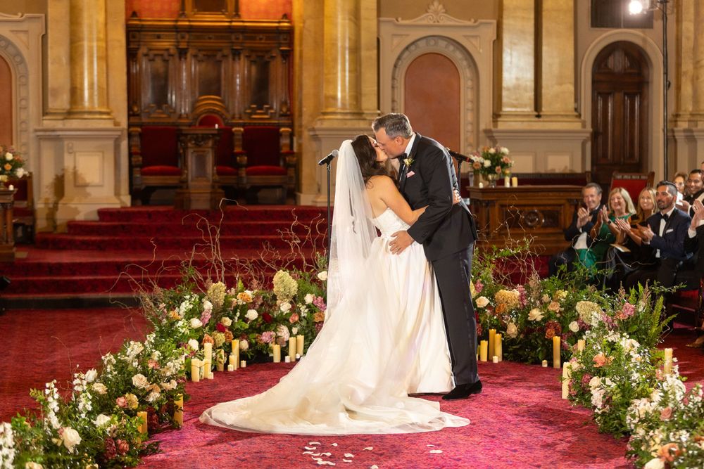 Newlyweds embrace during their ceremony in an ornate church decorated with floral arrangements.
