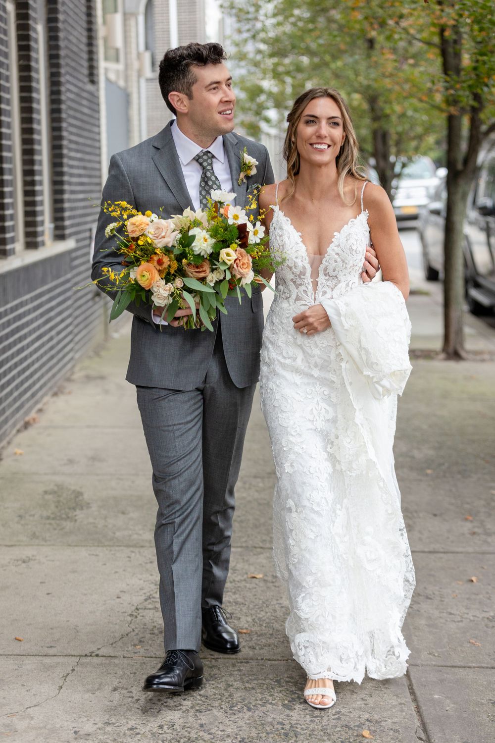 A bride in a lace gown and groom in a gray suit walk together on a sidewalk while carrying a vibrant fall bouquet.