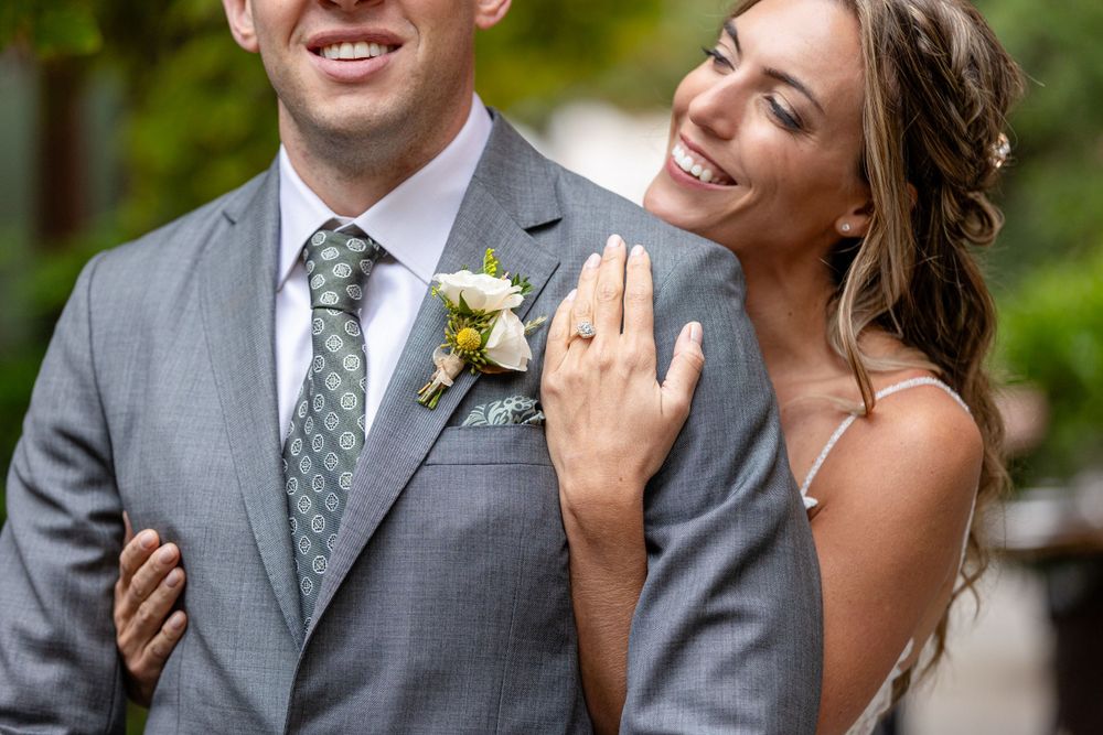 A joyful wedding couple shares a candid moment outdoors, with the groom wearing a grey suit and floral boutonniere.