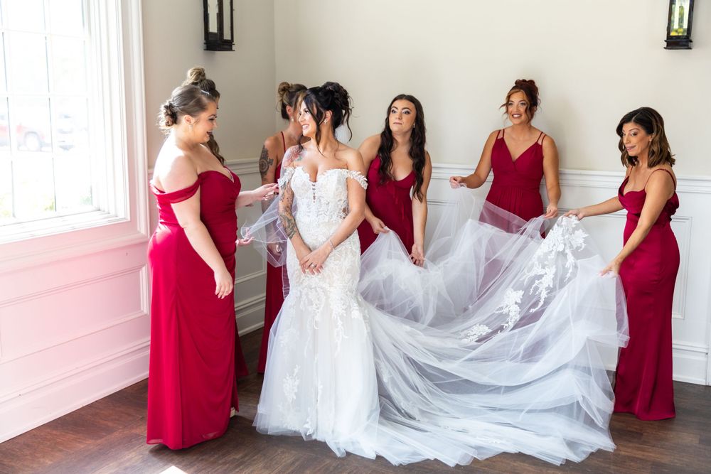 Bridesmaids in red dresses help adjust a white wedding gown with flowing tulle train in a bright indoor setting.
