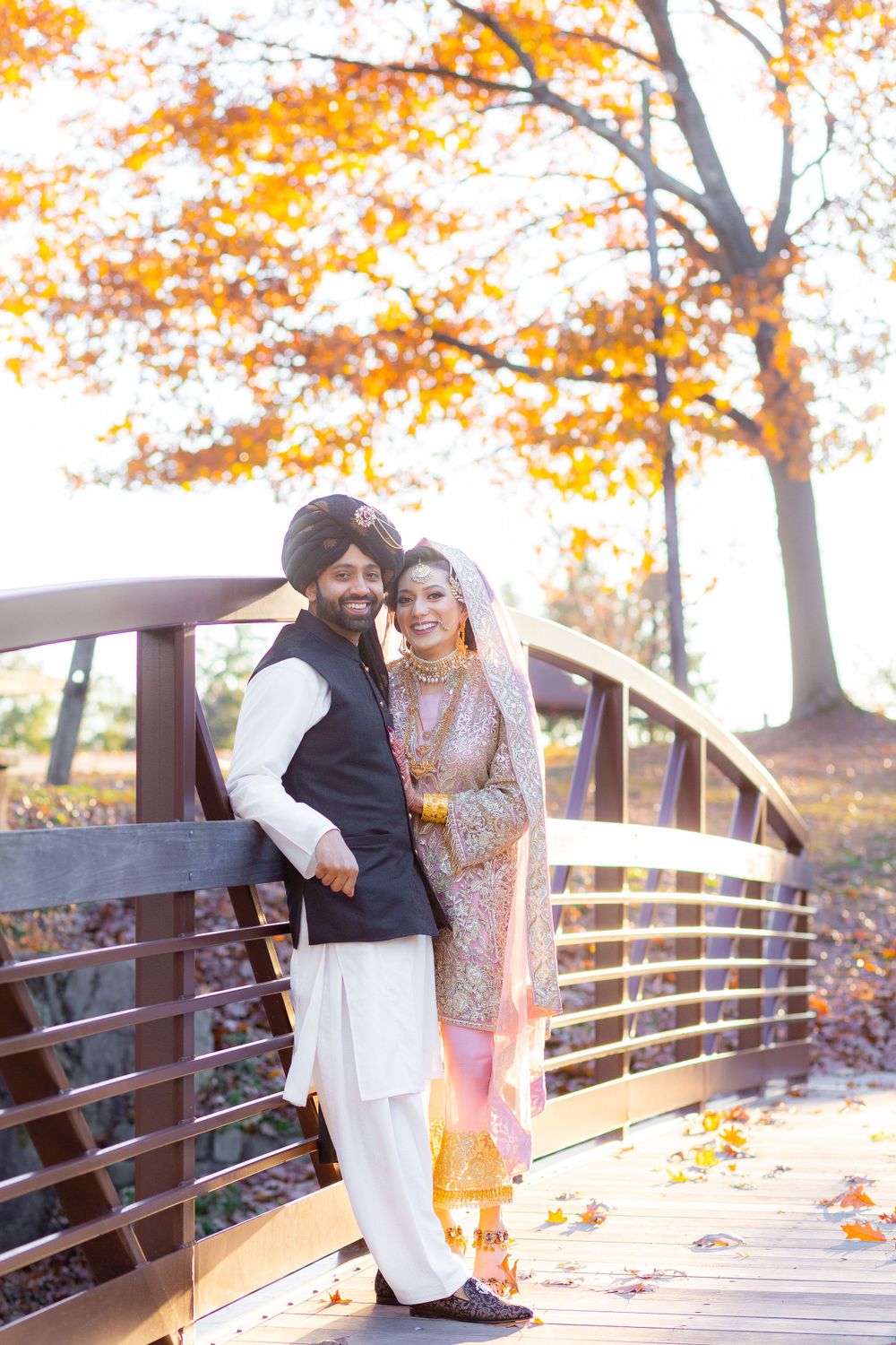 Couple in traditional Indian attire pose on autumn bridge with orange foliage.