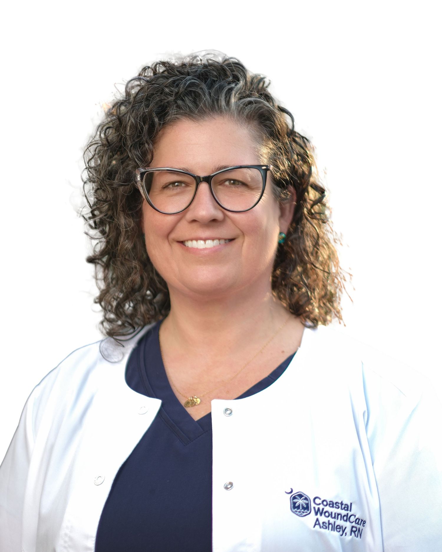 Healthcare professional in a white lab coat and glasses smiling while standing outdoors with trees in background.