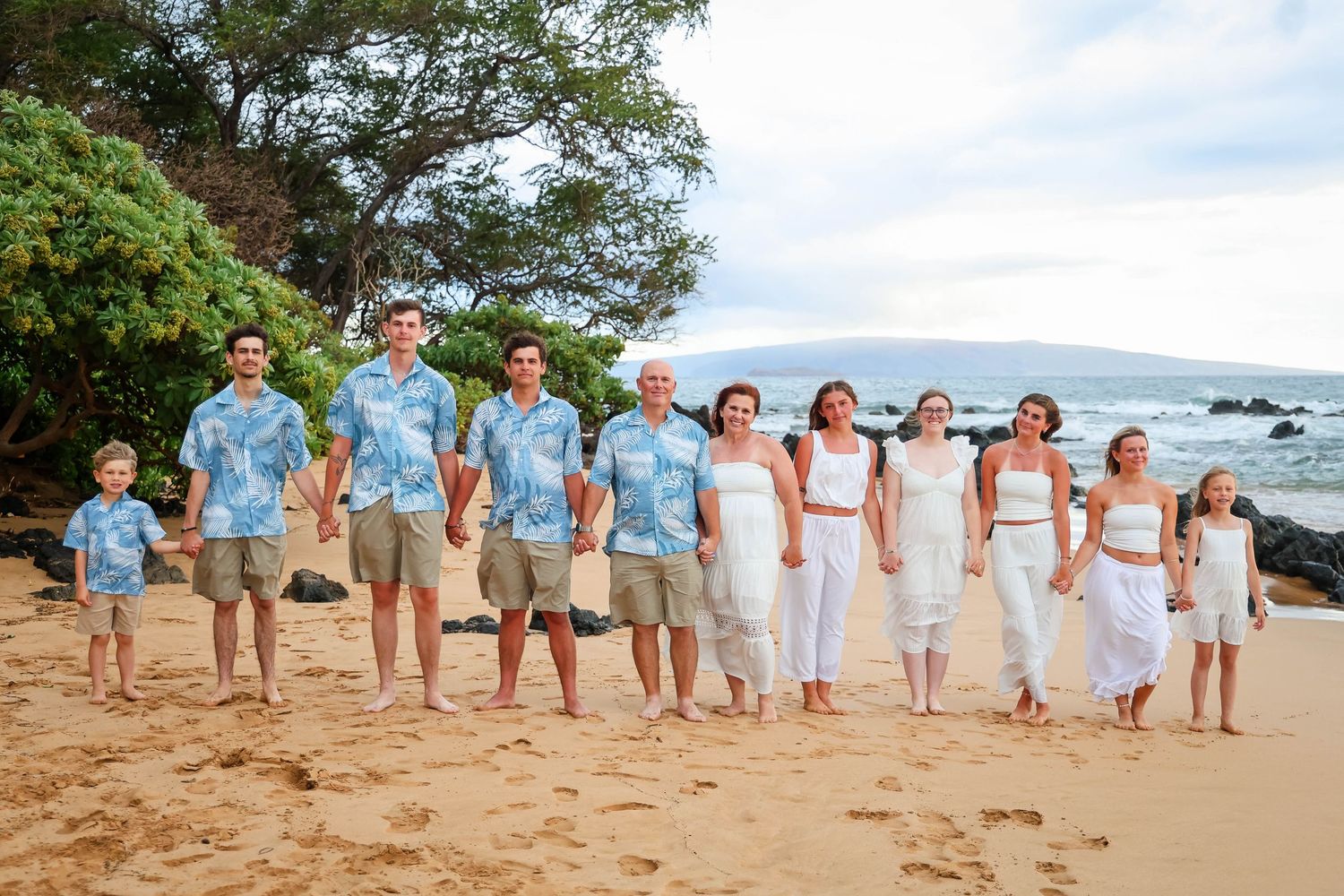 A group of people in coordinated white and blue outfits pose together on a scenic beach at sunset.