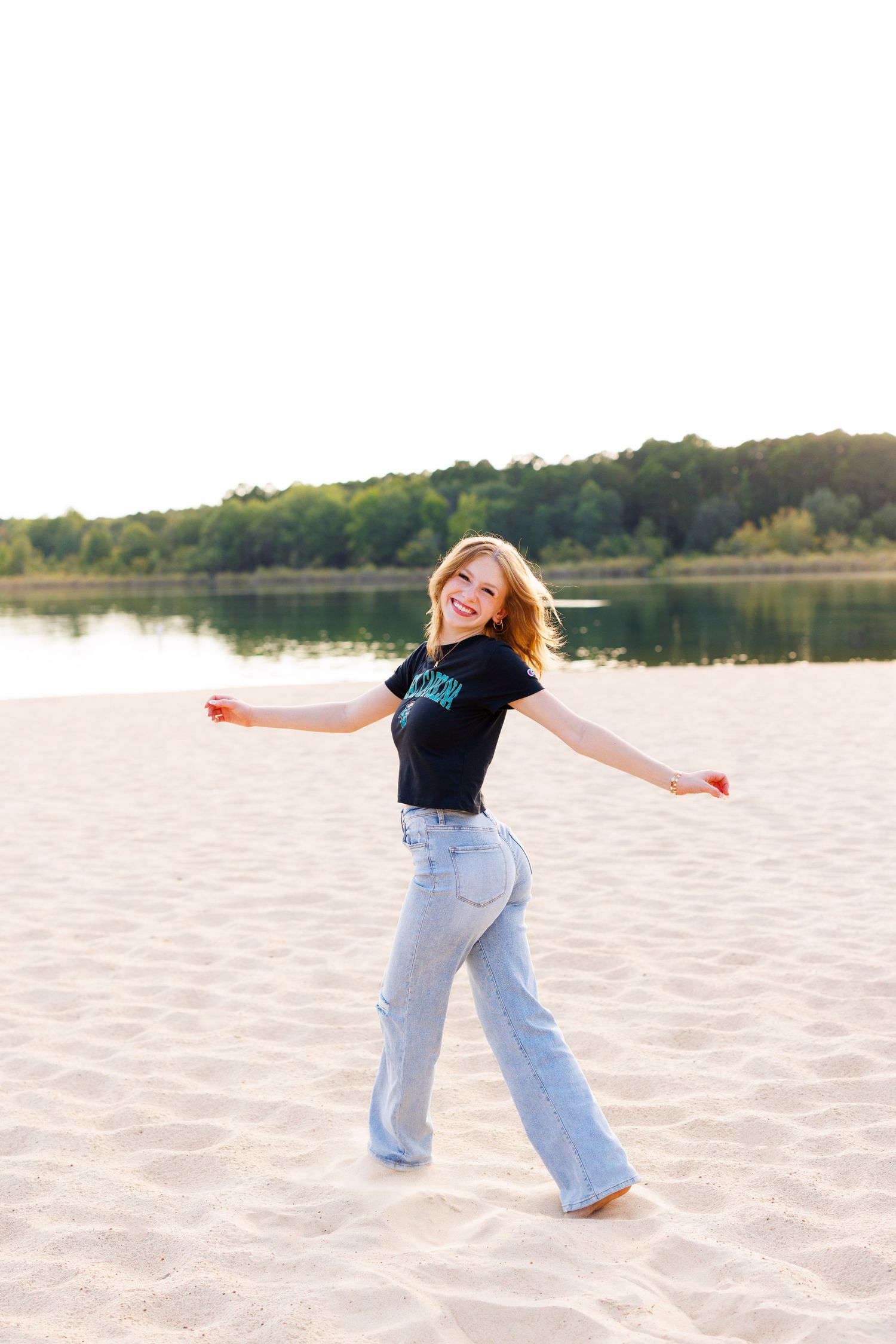 A carefree moment captured on a sandy beach with light blue jeans and black top against a lake backdrop at sunset.