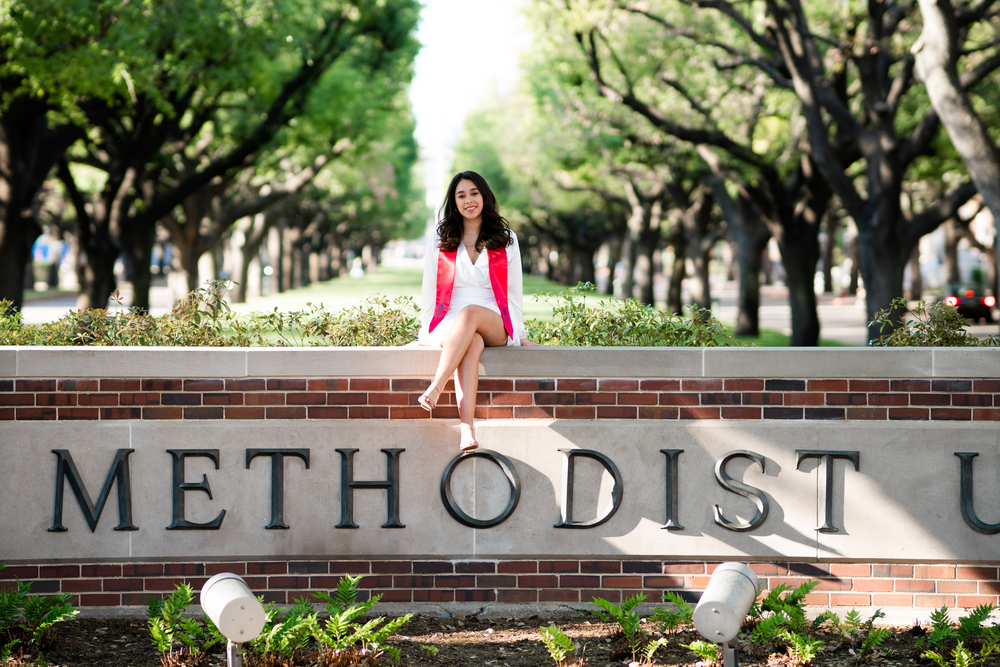 Graduate in white dress and pink blazer sits on brick wall with Methodist U sign and tree-lined path.