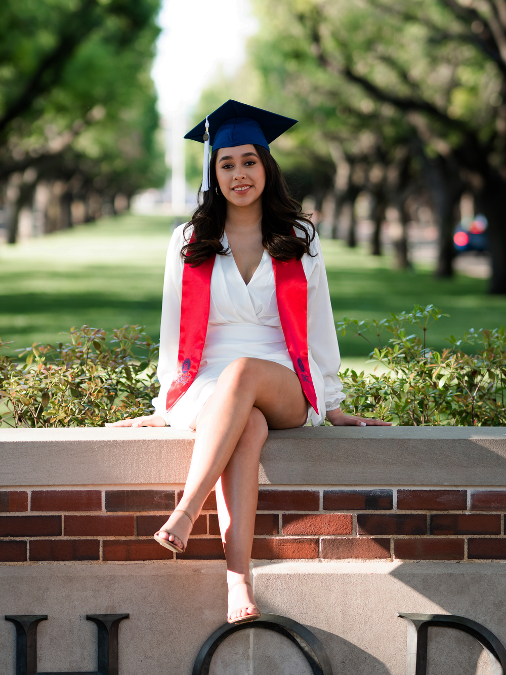 Graduate in cap and gown sits elegantly on brick wall during graduation photoshoot.