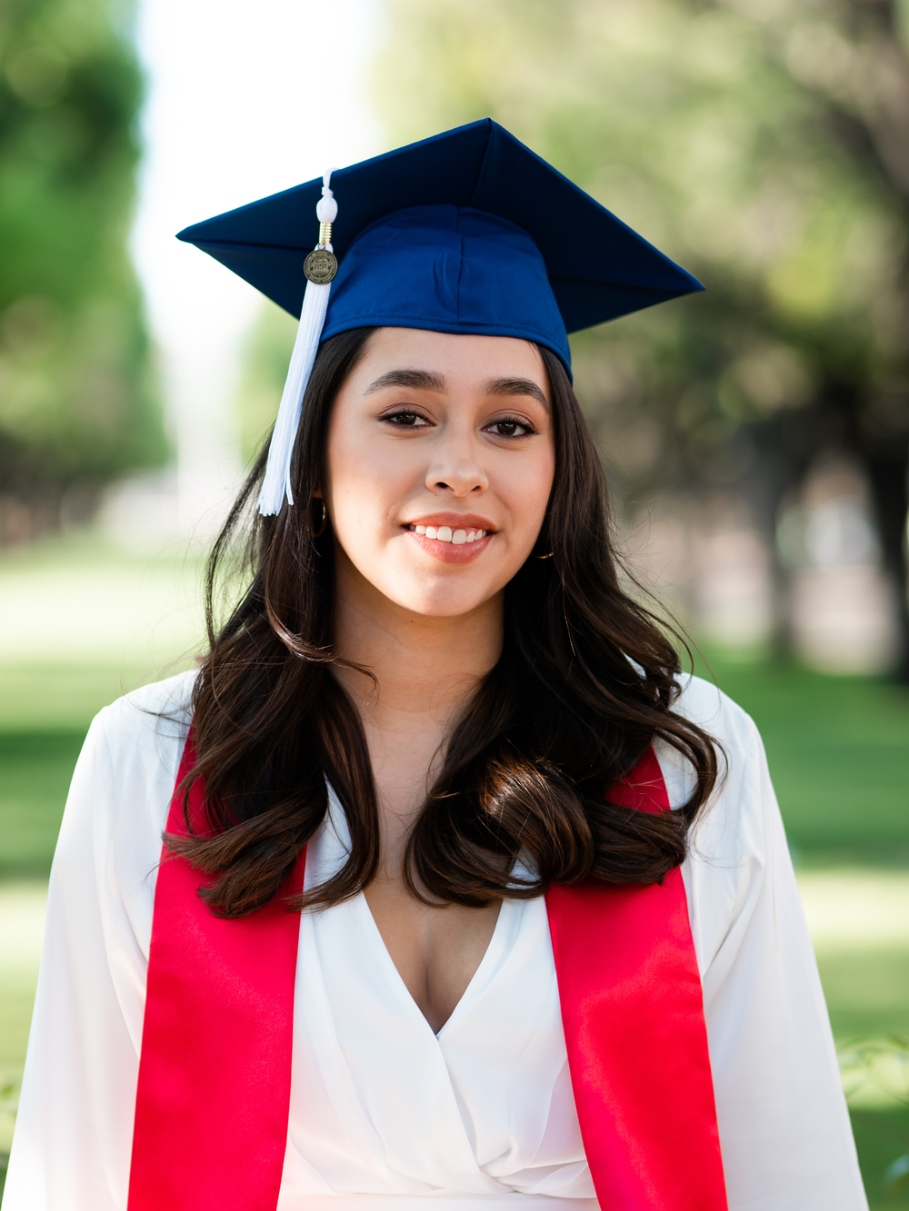 Graduate wearing blue cap with white tassel and red stole poses in white graduation gown.