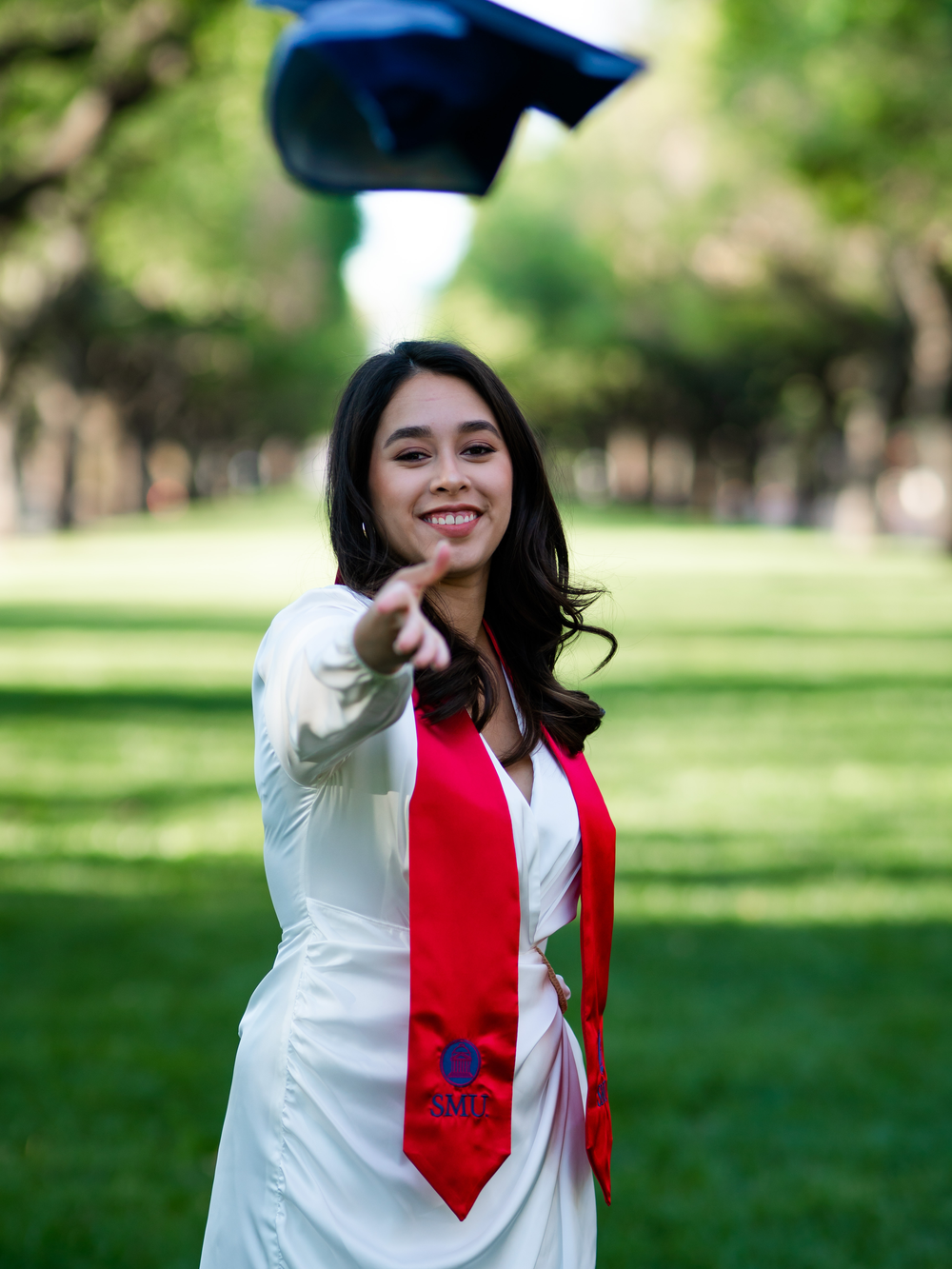 Graduate in cap and gown with red stole points playfully at camera outdoors.
