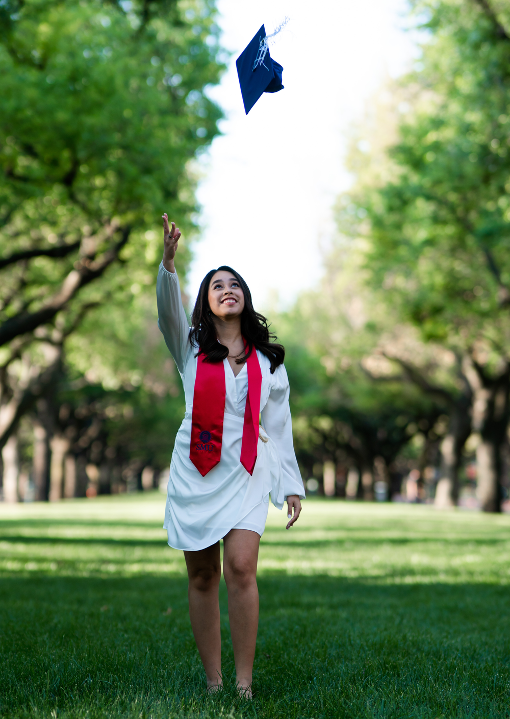 Graduate in white dress and red stole throws blue cap in air on tree-lined campus lawn.