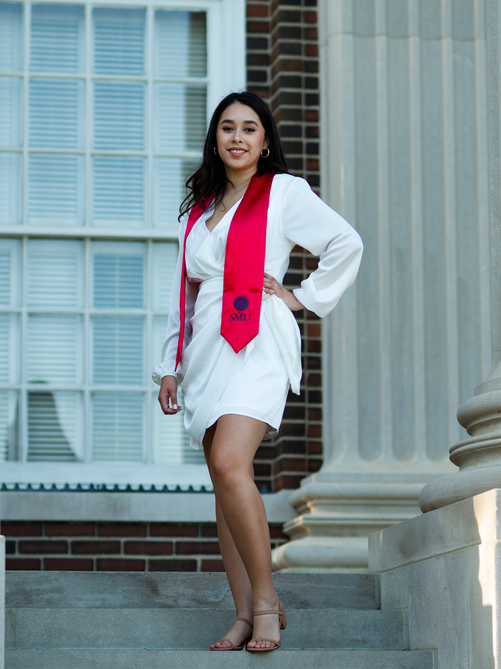 Graduate in white dress and red stole poses confidently on stone steps beside classical columns.