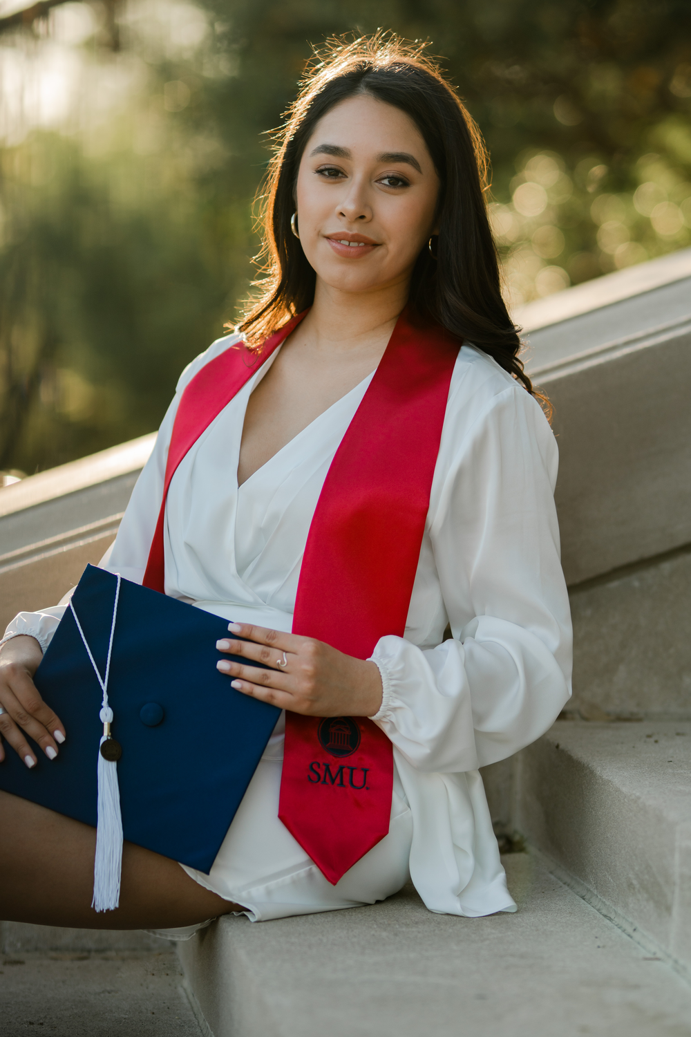 Graduate in white dress holds blue diploma folder while sitting on concrete steps outdoors.