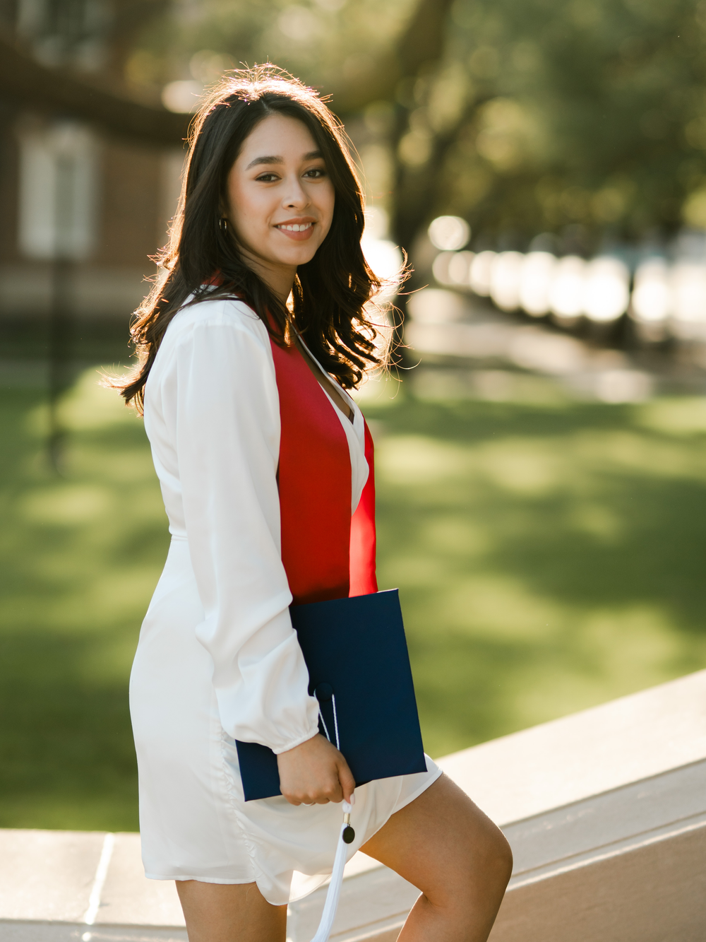 Graduate in white dress with red stole holds diploma on sunny campus walkway.