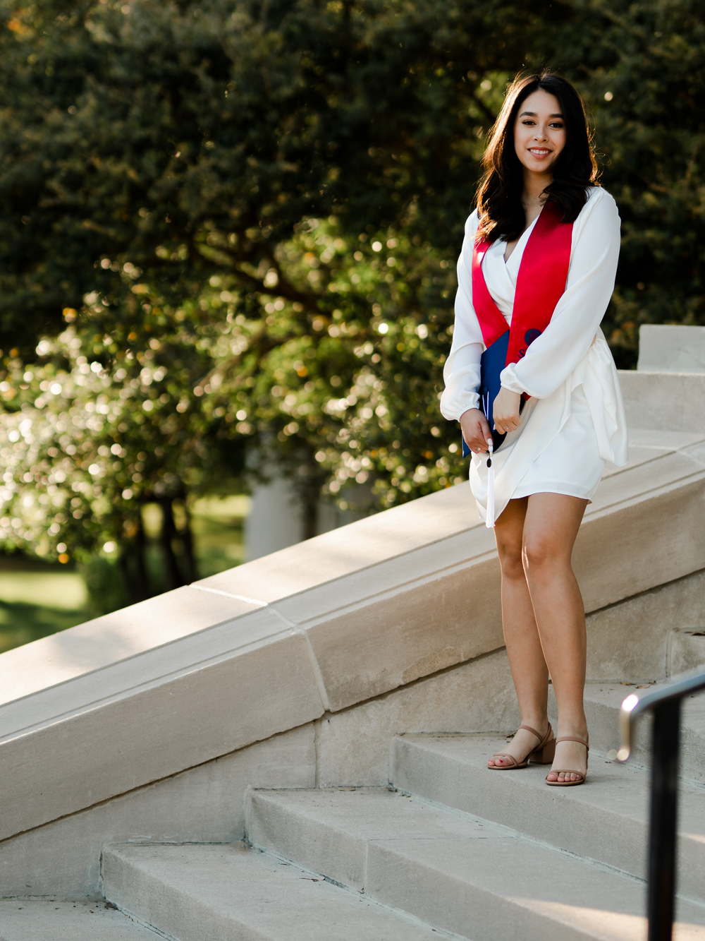 Graduate in white dress and red stole poses on stone steps during golden hour.