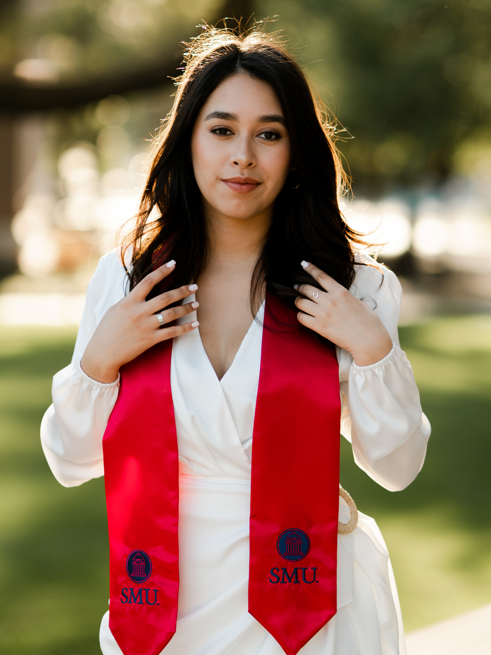 Graduate wearing white dress and red SMU graduation stole poses in outdoor setting.